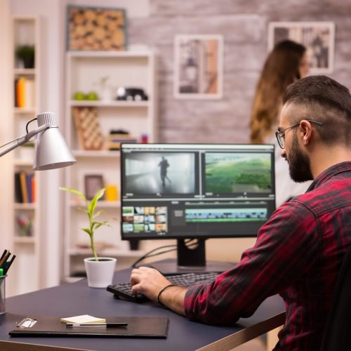 Back view of creative filmmaker working on a movie on laptop. Girlfriend in the background.